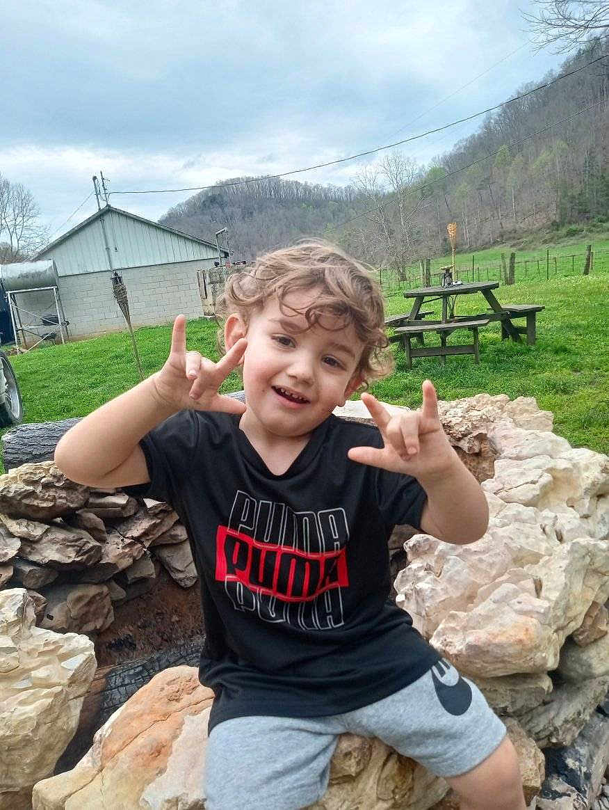 Rebel is registered to the contest to win money with this photo: child, boy, curly_hair, smiling, rock, outdoor, grass, picnic_table, cloudy_sky, hill, casual_clothing, black_tshirt, shorts, playful, nature, fence, wooden_bench, daylight, hand_sign, happy