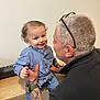 adult, arm, child, face, glasses, hair, hand, happy, holding, indoor, interaction, jeans, person, shirt, smile, table, tattoo, toddler, watch, wood