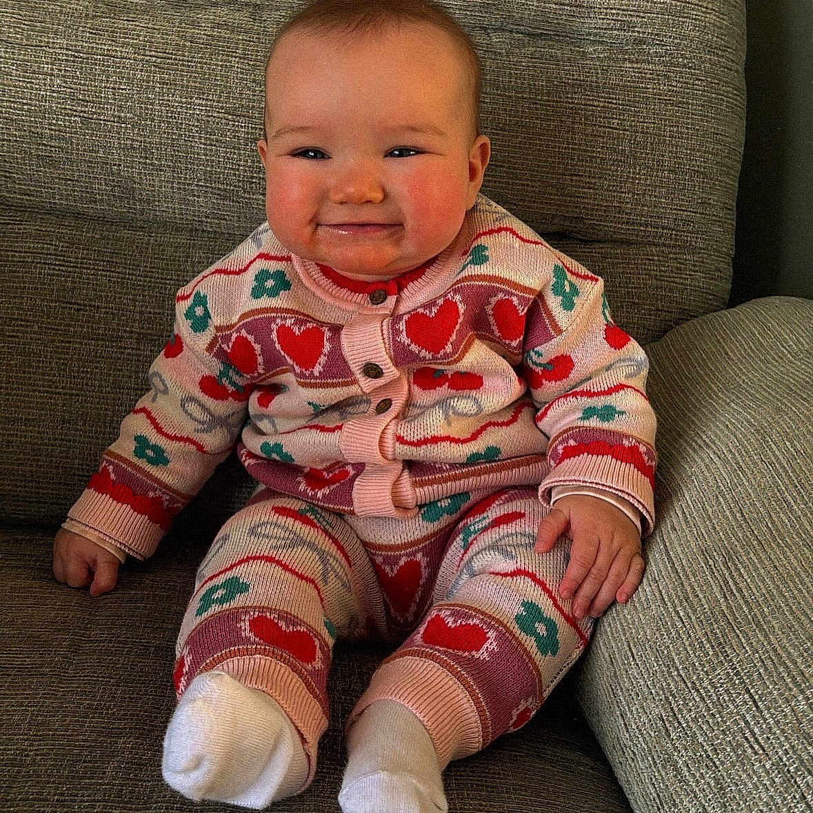 Enya is registered to the contest to win money with this photo: baby, child, couch, cozy, cute, face, floral_pattern, hand, happy, heart_pattern, indoor, infant, knitted_clothing, portrait, sitting, smiling, sock, texture, warm_clothing, young_child