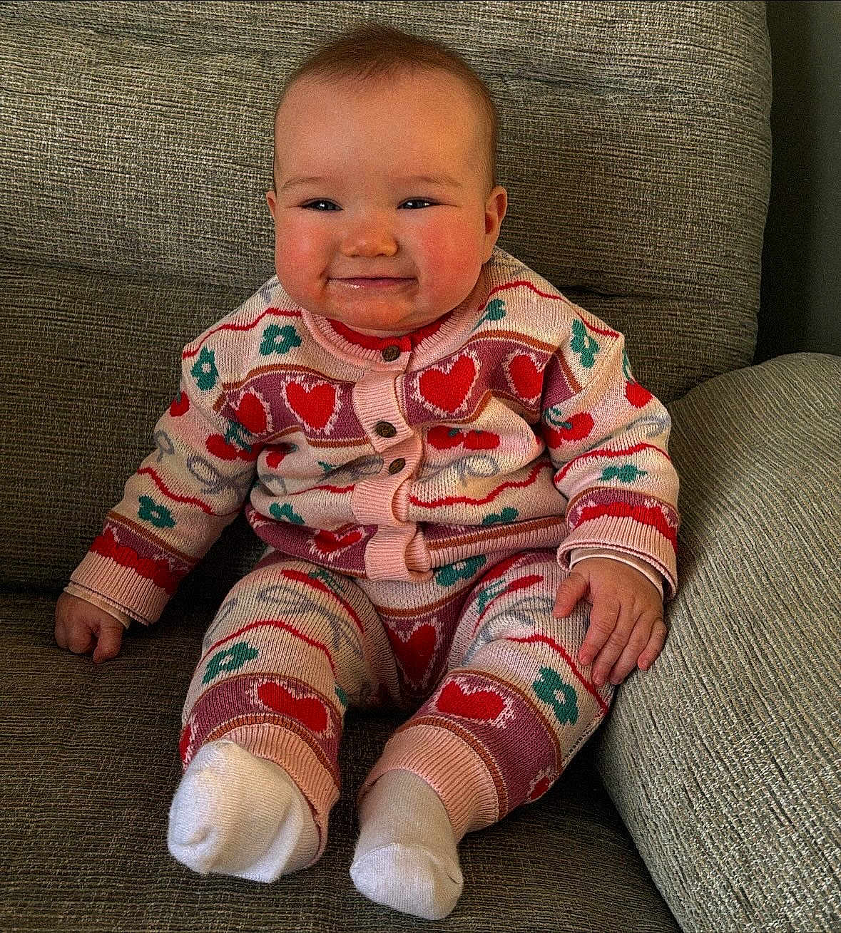 Enya is registered to the contest to win money with this photo: baby, child, smiling, sitting, couch, knitted_clothing, heart_pattern, floral_pattern, sock, cute, indoor, portrait, cozy, infant, happy, young_child, face, hand, warm_clothing, texture