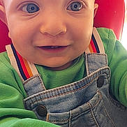 Natan participe au concours pour gagner de l'argent avec cette photo : baby, child, smiling, blue_eyes, denim_overalls, green_shirt, rainbow_straps, closeup, portrait, happy, cute, infant, young_child, hand, face, sitting, red_background, clothing, person, indoors