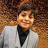 black_clothing, boy, bronze, child, dress_shirt, facial_expression, fashion, formalwear, gray_clothing, indoor, looking_at_camera, metallic_background, photography, portrait, short_hair, smiling, standing, suit, textured_wall, vest