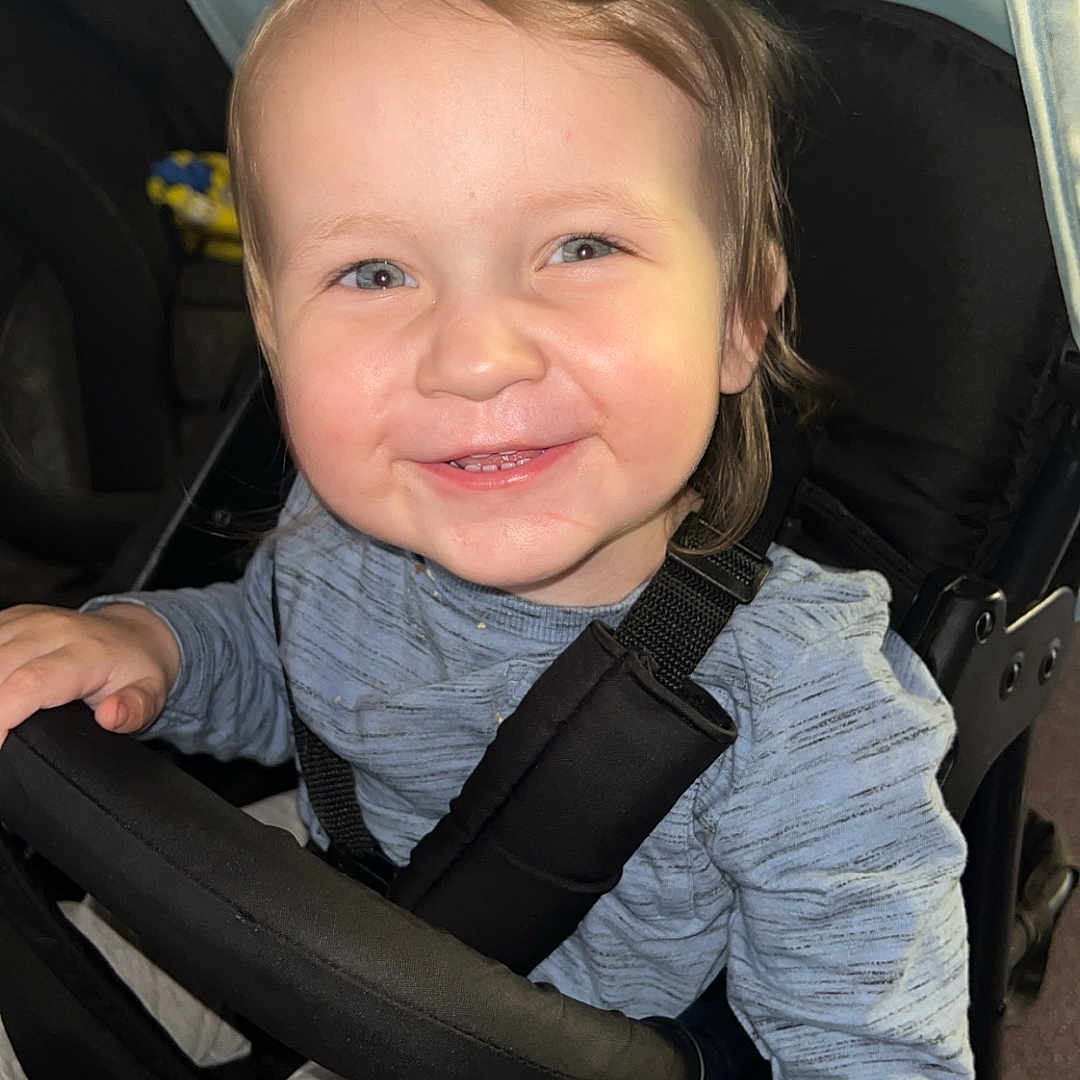 Maverick is registered to the contest to win money with this photo: accessories, baby, belt, bodypart, clothing, coat, face, finger, hand, happy, head, jacket, pants, person, photography, portrait, smile, stroller, transportation, vehicle