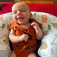 Colton is registered to the contest to win money with this photo: baby, infant, smiling, orange_onesie, baby_seat, bear_pattern, tree_pattern, cushion, red_blanket, wooden_drawer, indoor, happy, cute, child, person, portrait, sitting, home, clothing, face