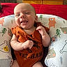 baby, infant, smiling, orange_onesie, baby_seat, bear_pattern, tree_pattern, cushion, red_blanket, wooden_drawer, indoor, happy, cute, child, person, portrait, sitting, home, clothing, face