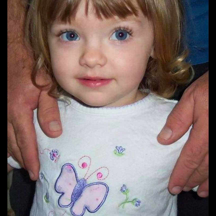 Vera is registered to the contest to win money with this photo: adult_hands, blue_eyes, butterfly_design, casual_clothing, child, childhood, close_up, curly_hair, cute, hands, human, indoor, looking_up, person, portrait, smiling, standing, toddler, white_shirt, young_child