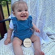 Iyla is registered to the contest to win money with this photo: baby, infant, smile, blue_romper, white_bow, crocheted_blanket, outdoor, grass, wooden_sign, happy, child, cute, milestone, portrait, sitting, person, toes, hand, face, young_child