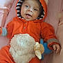 baby, infant, child, onesie, orange_clothing, hood, ears, plush_toy, pink_background, smiling, tongue_out, cute, soft_texture, lying_down, cozy, playful, happy, person, toddler, indoor