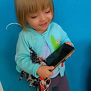 Mélody participe au concours pour gagner de l'argent avec cette photo : child, girl, blue_door, cardigan, scarf, phone, black_shoes, floor_tiles, standing, smiling, indoors, short_hair, casual_clothing, playful, happy, person, full_body, wall, footwear, young_child