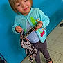 Mélody participe au concours pour gagner de l'argent avec cette photo : toddler, child, blue_wall, cardigan, leggings, black_shoes, floor_tiles, indoor, holding, book, patterned_cloth, short_hair, cute, smiling, standing, person, footwear, floor, wall, curious