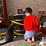 bare_feet, boy, cabinet, carpet, cat, child, gray_cat, green_ball, hot_wheels, indoor, kneeling, pet, playing, plush_toy, red_shirt, scattered_toys, textured_wall, toddler, toy_track, toys
