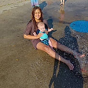 Ember is registered to the contest to win money with this photo: child, baby, water, splash_park, wet_ground, playing, summer, outdoor, barefoot, sitting, holding, fun, water_spray, shadow, peace_sign, casual_clothing, smiling, sunny_day, friends, park