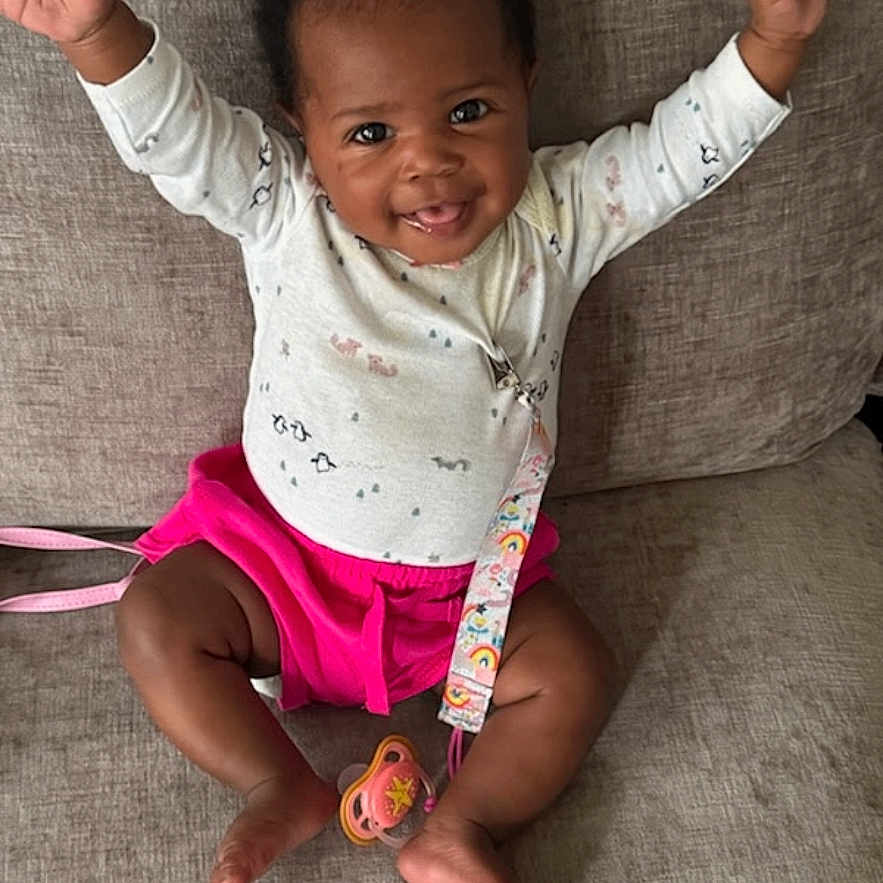 Ariana Durodola joined the competition — help win amazing prizes! arms_up, baby, black_hair, child, clothing, couch, cute, expression, face, happy, home, indoors, infant, joy, pacifier, person, pink_shorts, sitting, smiling, white_shirt