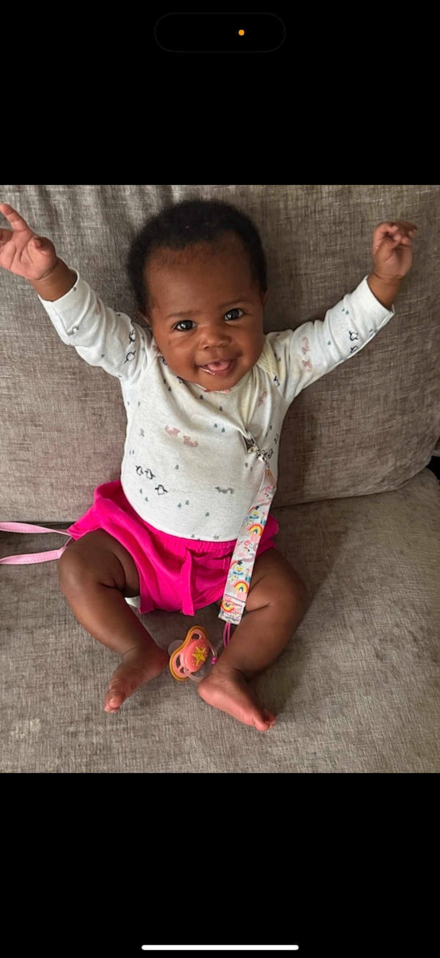 Ariana Durodola joined the competition — help win amazing prizes! arms_up, baby, black_hair, child, clothing, couch, cute, expression, face, happy, home, indoors, infant, joy, pacifier, person, pink_shorts, sitting, smiling, white_shirt