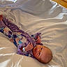 Catalina is registered to the contest to win money with this photo: baby, bed, bedroom, child, cute, eyes_open, face, feet, floral_pattern, hands, indoors, infant, lying_down, photography, portrait, purple_clothing, relaxed, satin, soft_lighting, white_bedding