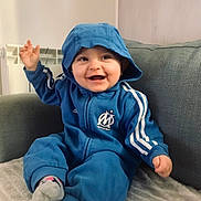 Giulia participe au concours pour gagner de l'argent avec cette photo : baby, child, smiling, blue_hoodie, sitting, couch, indoor, happy, cozy, cute, person, soft_lighting, hood, casual_clothing, sock, furniture, portrait, young_child, playful, warm