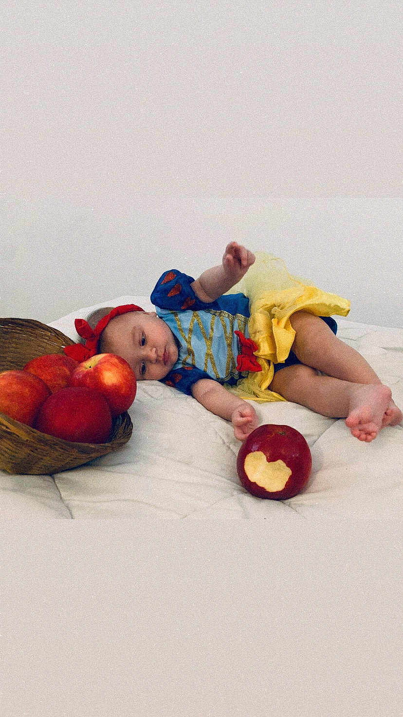 Giulia participe au concours pour gagner de l'argent avec cette photo : baby, child, infant, apple, basket, bite, costume, dress, yellow_skirt, blue_top, red_bow, lying_down, white_background, soft_surface, fruit, hand, foot, cute, indoor, portrait