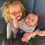 Kandie is registered to the contest to win money with this photo: child, baby, couch, curly_hair, bib, smiling, happy, indoor, holding, clothing, person, face, sitting, window, cushion, cute, family, portrait, young_child, toddler