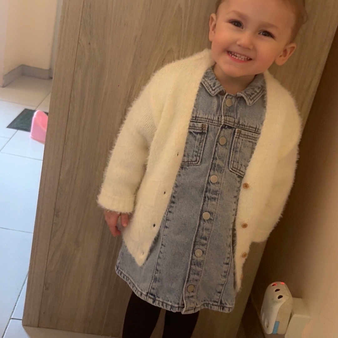 Eliyanah a rejoint le concours — aidez-le/la à gagner de superbes lots ! child, clothing, cute, denim_dress, hair_topknot, hallway, happy, indoor, pink_sneakers, portrait, power_plug, shoes, small_child, smiling, standing, tile_floor, toddler, wall_socket, white_cardigan, wood_panel