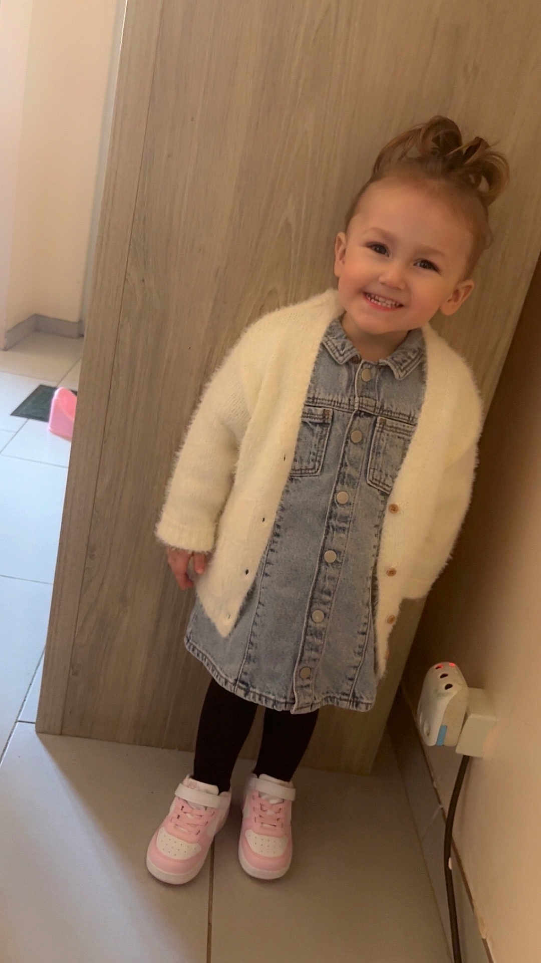 Eliyanah a rejoint le concours — aidez-le/la à gagner de superbes lots ! child, toddler, smiling, denim_dress, white_cardigan, pink_sneakers, hair_topknot, standing, indoor, wood_panel, tile_floor, wall_socket, power_plug, happy, portrait, cute, clothing, small_child, shoes, hallway