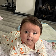 Aria is registered to the contest to win money with this photo: baby, infant, onesie, pumpkin_pattern, blanket, floor, fireplace, shelf, indoors, curious, lying_down, person, child, face, head, hand, home, wood_floor, cozy, cute