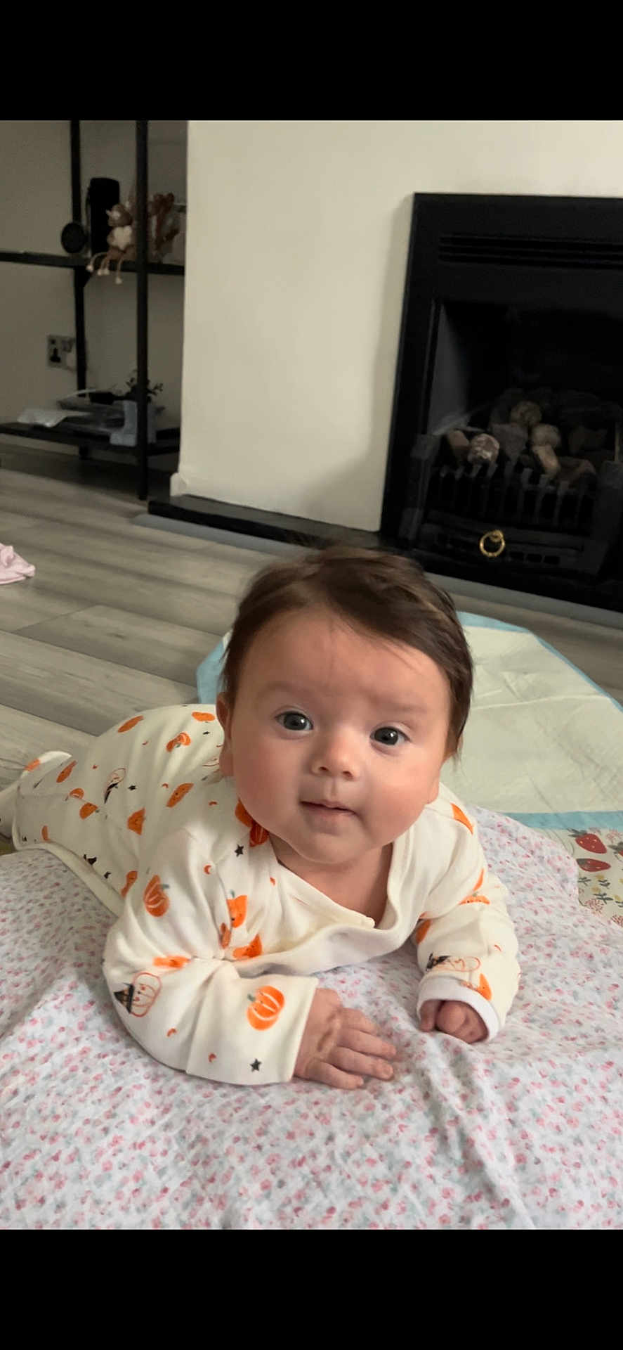 Aria is registered to the contest to win money with this photo: baby, infant, onesie, pumpkin_pattern, blanket, floor, fireplace, shelf, indoors, curious, lying_down, person, child, face, head, hand, home, wood_floor, cozy, cute