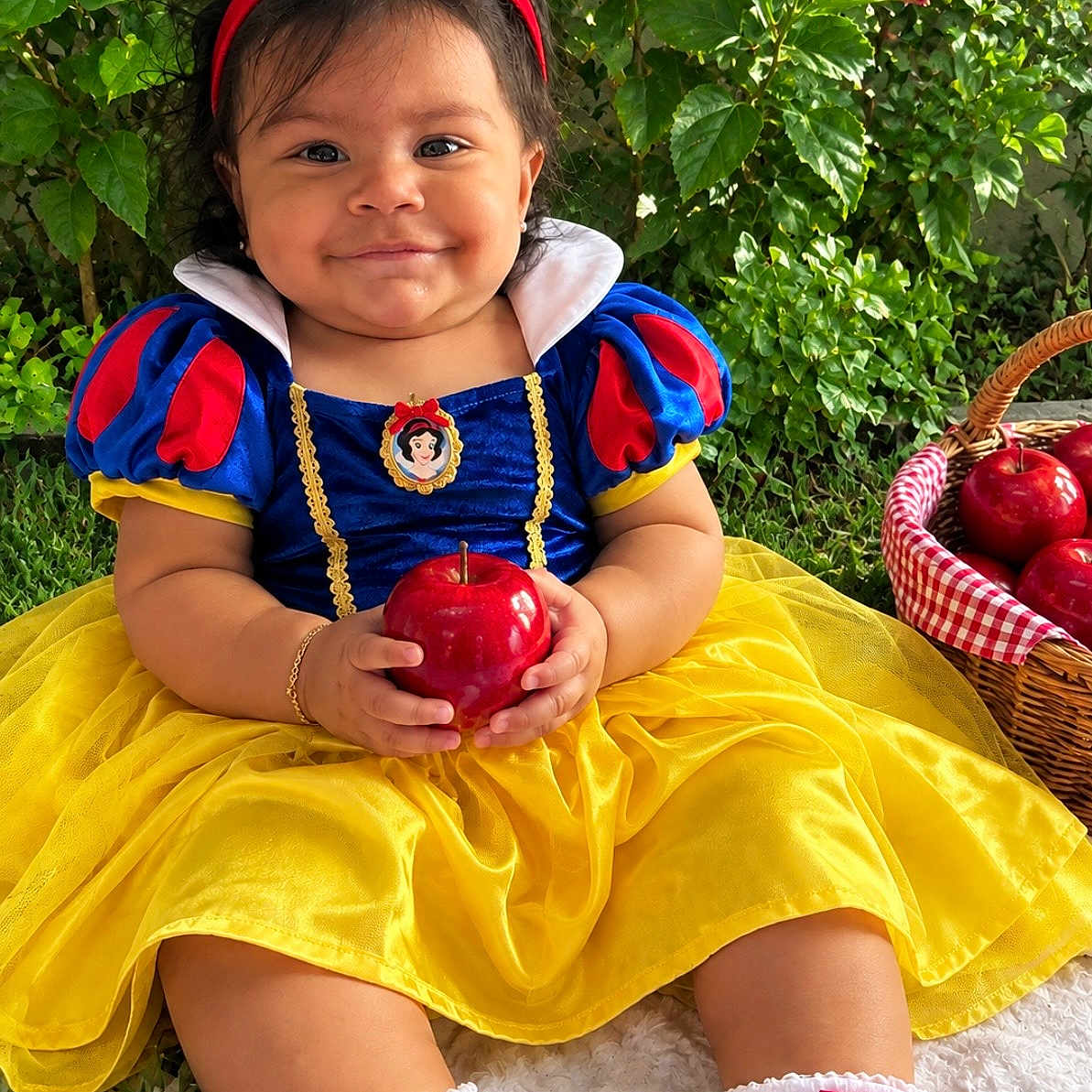 Zara Isabella joined the competition — help win amazing prizes! baby, child, toddler, portrait, smiling, happy, snow_white_costume, red_bow, blue_dress, yellow_skirt, apple, fruit_basket, outdoor, garden, greenery, grass, storybook, princess, sitting, family_friendly