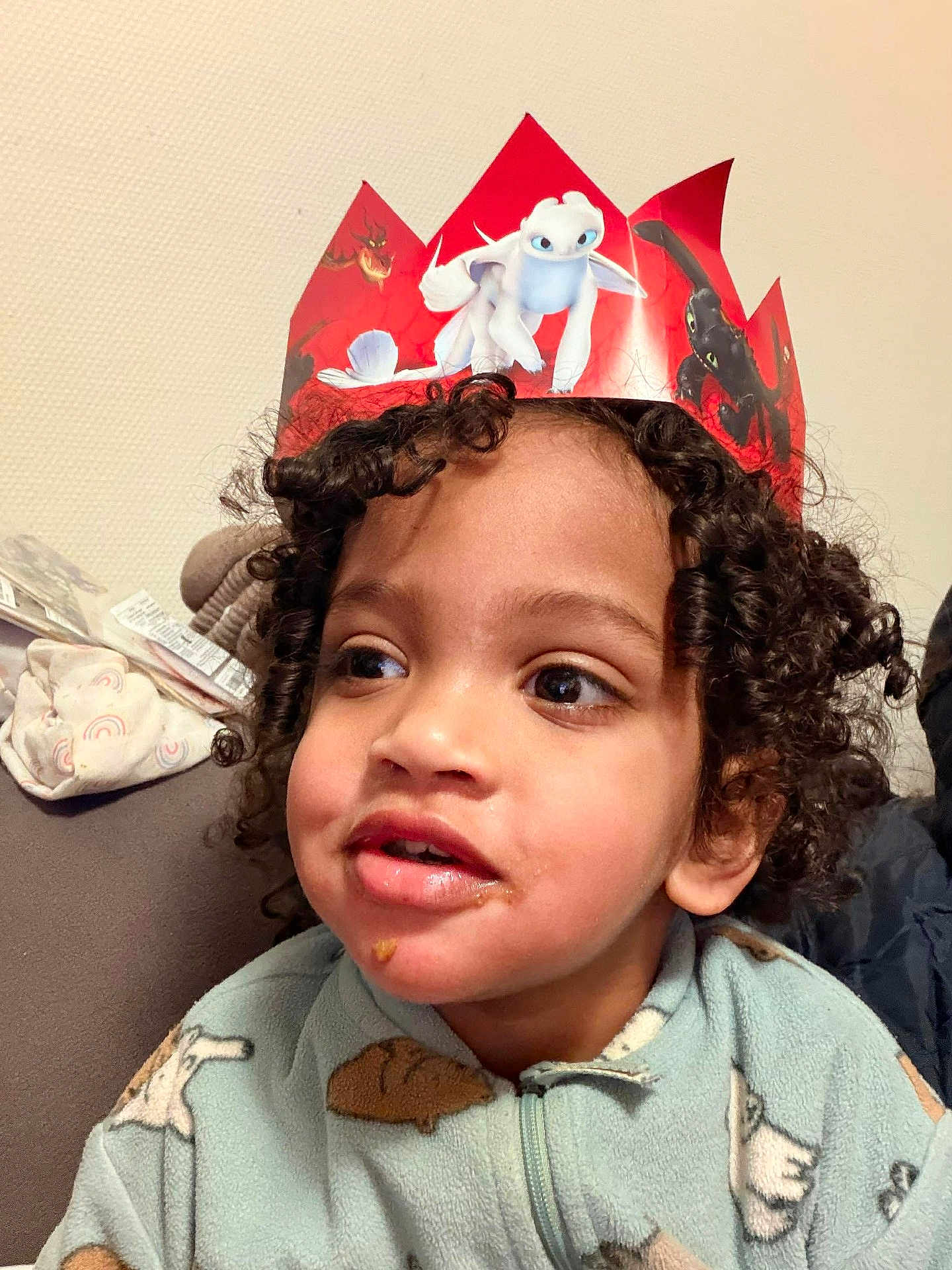 Lawson Frontin participe au concours pour gagner de l'argent avec cette photo : child, curly_hair, crown, red_crown, paper_crown, dragon, face, indoors, cozy, clothing, onesie, animal_pattern, food_on_face, couch, furniture, smiling, portrait, person, young_child, happy