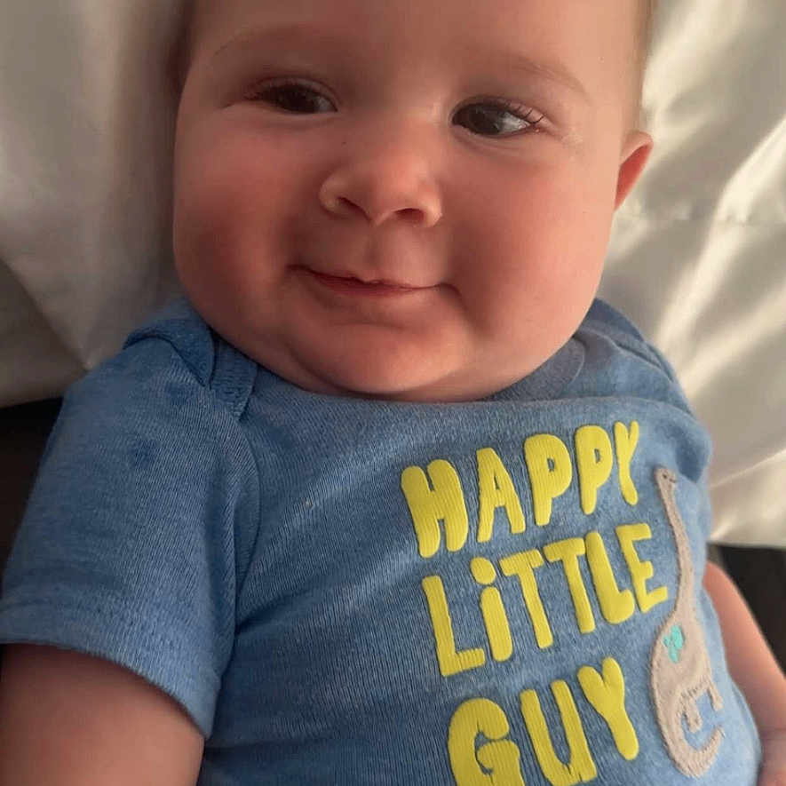 Brextyn is registered to the contest to win money with this photo: baby, blue_shirt, cheeks, child, close_up, cute, face, happy, head, indoors, infant, lying_down, person, pillow, short_hair, skin, smiling, text_on_clothing, toddler, young_child