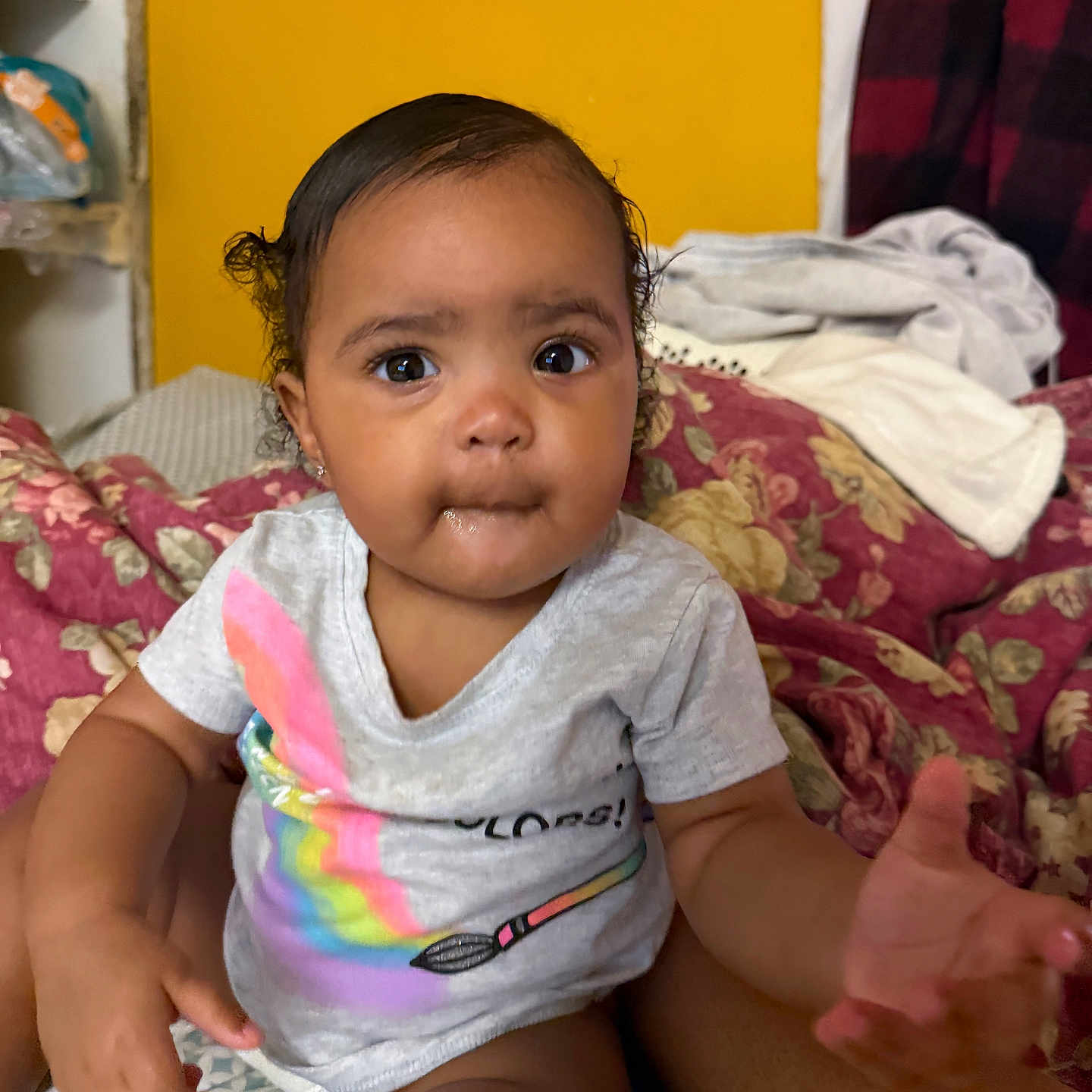 Khalaya is registered to the contest to win money with this photo: baby, child, infant, portrait, sitting, bed, bedroom, floral_bedding, yellow_wall, rainbow_shirt, gray_shirt, curly_hair, pigtails, brown_eyes, wide_eyed, curious, indoors, home, person, smiling