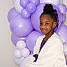 background, balloons, bow, celebration, cheerful, child, decor, dress, girl, hair_bun, indoor, jacket, looking_at_camera, party, portrait, purple, sitting, smile, studio_lighting, white_jacket
