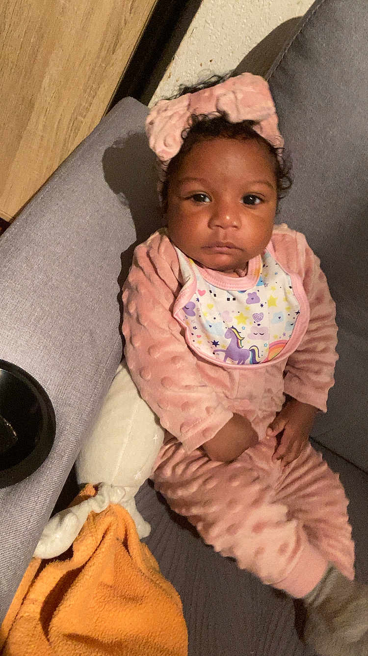 Luisa participe au concours pour gagner de l'argent avec cette photo : baby, bib, blanket, child, couch, curious_expression, dark_eyes, hand, headband, home, indoor, infant, pillow, pink_outfit, plush_toy, portrait, relaxed, sitting, textured_fabric, warm_lighting