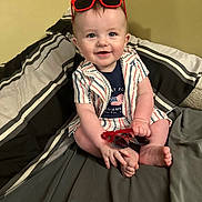 Cove is registered to the contest to win money with this photo: baby, bed, blanket, blue_eyes, casual_clothing, child, cute, foot, hand, happy, indoor, infant, patterned_clothing, person, red_sunglasses, short_hair, sitting, smile, striped_clothing, sunglasses