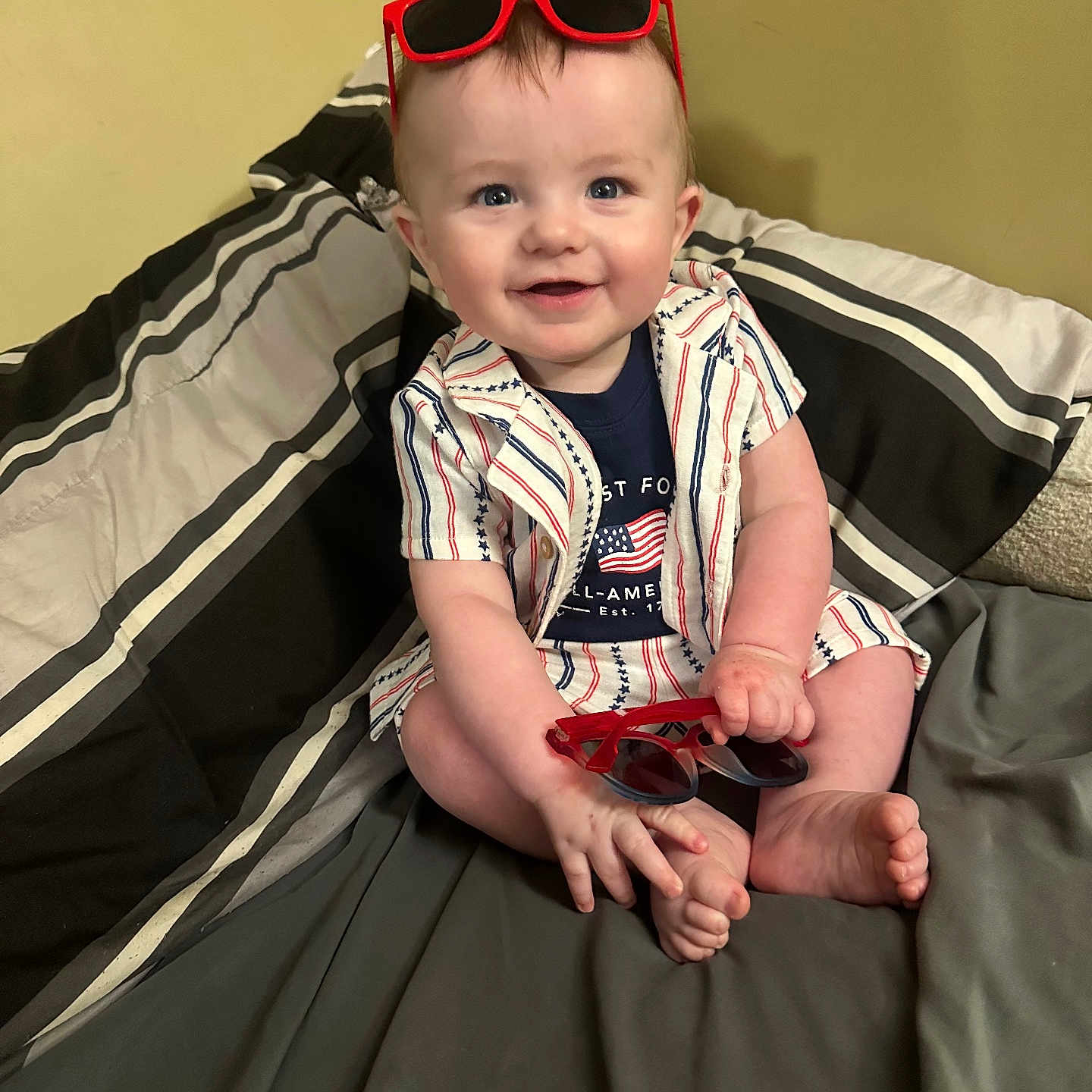 Cove is registered to the contest to win money with this photo: baby, bed, blanket, blue_eyes, casual_clothing, child, cute, foot, hand, happy, indoor, infant, patterned_clothing, person, red_sunglasses, short_hair, sitting, smile, striped_clothing, sunglasses