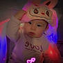 adorable, baby, bunny_hat, child, colorful_lights, cozy_clothing, cute, dim_lighting, expression, face, glowing_lights, hearts, indoors, infant, love_text, pink_hat, portrait, sitting, toy, young_child