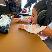 Arliss Selah is registered to the contest to win money with this photo: baby, pacifier, headphones, computer, keyboard, mouse, microphone, desk, hand, person, technology, indoor, monitor, wearing, clothing, cute, infant, electronics, reflection, window