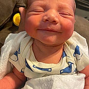Easteynn is registered to the contest to win money with this photo: baby, newborn, smiling, face, blanket, onesie, blue_pattern, white_clothing, infant, resting, couch, soft_texture, skin, hand, comfort, closeup, child, indoors, peaceful, sleepy