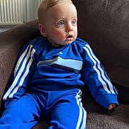 Lucas is registered to the contest to win money with this photo: toddler, child, blue_tracksuit, sitting, couch, indoor, white_shoes, curious_expression, young_child, face, person, home, casual_clothing, cozy, light, window, door, furniture, sock, portrait