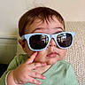 baby, child, sunglasses, green_clothing, hand, face, reflection, knitted_couch, indoor, portrait, cute, thoughtful, casual, seated, eyewear, young_child, skin, expression, fingers, hair