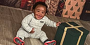 Kimani Mason is registered to the contest to win money with this photo: baby, child, gift_box, christmas_tree, presents, flooring, holiday, smiling, indoor, festive, decorations, red, green, gold, wrapped_gifts, happy, clothing, sitting, celebration, home
