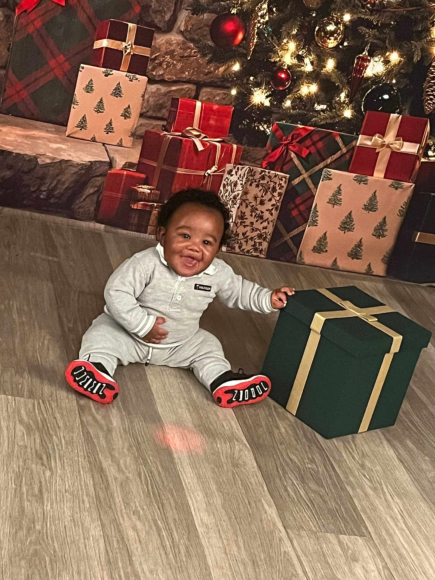 Kimani Mason is registered to the contest to win money with this photo: baby, child, gift_box, christmas_tree, presents, flooring, holiday, smiling, indoor, festive, decorations, red, green, gold, wrapped_gifts, happy, clothing, sitting, celebration, home