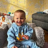 baby, child, blue_outfit, smiling, sitting, indoor, fluffy_chair, toy, wallpaper, sunlight, shadow, couch, floor, happy, person, head, face, sock, cozy, cartoon_character