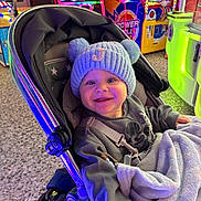 Jamie is registered to the contest to win money with this photo: baby, stroller, hat, pom_poms, sweatshirt, blanket, indoor, arcade, neon_lights, smile, child, face, happy, toy, seat, floor, colorful, game_machine, person, playful