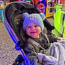 baby, stroller, hat, pom_poms, sweatshirt, blanket, indoor, arcade, neon_lights, smile, child, face, happy, toy, seat, floor, colorful, game_machine, person, playful