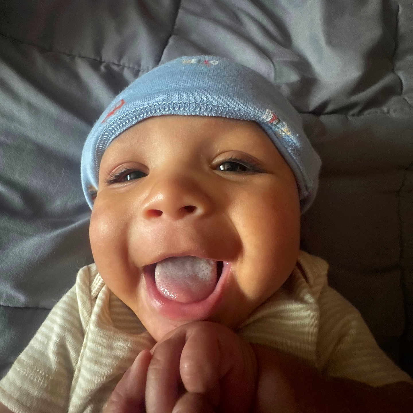 Julien joined the competition — help win amazing prizes! baby, infant, smiling, tongue_out, blue_cap, holding_hands, closeup, face, cheeks, cute, child, lying_down, blanket, soft_lighting, happy, expressive, person, portrait, indoors, skin