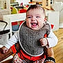 baby, smiling, indoor, bib, clothing, person, happy, child, holding, furniture, floor, wooden_floor, background, adult_hand, sitting, casual, cute, infant, expression, joy