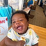baby, smiling, curly_hair, onesie, indoor, person, happy, child, face, cute, patterned_clothing, table, adult, background, blurry, holding, joyful, expression, casual, restaurant