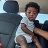 baby, child, infant, car_interior, car_seat, center_console, onesie, tattoo, arm, curly_hair, window, sky, seat_headrest, person, indoor, casual_clothing, young_child, looking, seated, supporting_hand