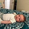 baby, infant, bed, blanket, pattern, clothing, white_shirt, grey_pants, socks, indoor, room, door, furniture, lying_down, face, person, calm, soft_lighting, home, portrait