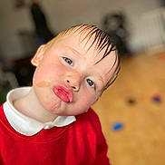 Archie is registered to the contest to win money with this photo: child, face, pout, wet_hair, red_sweater, white_collar, indoor, floor, toy, person, playful, cute, portrait, blurry_background, expression, skin, head, hair, young, funny_face